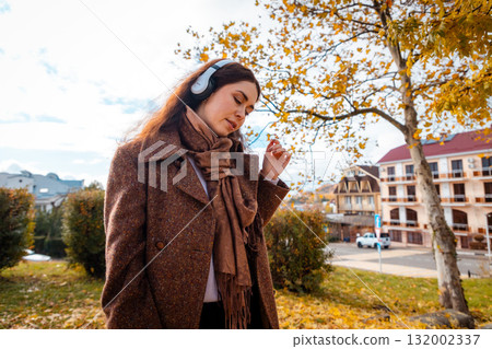 Mid shot of a happy young Caucasian pretty woman in a coat listening to music with headphones in park. Concept of happiness and carefree Mid shot of a happy young Caucasian pretty woman in a coat listening to music with headphones in park. Concept of happiness and carefree 132002337