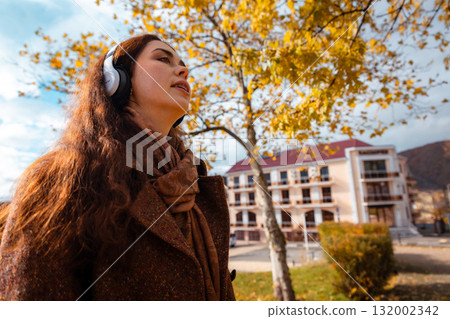 Bottom view of a Caucasian young woman in a coat listening to music with headphones in autumn park. Concept of happiness and carefree Bottom view of a Caucasian young woman in a coat listening to music with headphones in autumn park. Concept of happiness and carefree 132002342