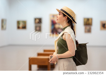 Side view of a young Caucasian woman in a straw hat and elegant clothes contemplating modern art in the gallery. Copy space. The concept of the Museum Day 132002354