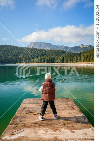 Vertical photo of boy standing on wooden pier and looking at Black Lake surrounded by pine forest and mountains in Durmitor National Park, Montenegro. Tranquil natural landscape with reflections on Vertical photo of boy standing on wooden pier and looking at Black Lake surrounded by pine forest and mountains in Durmitor National Park, Montenegro. Tranquil natural landscape with reflections on 132002654