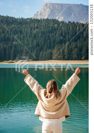 Half-length view of a woman standing on a wooden pier with raised arms, enjoying the scenic Black Lake and forest landscape in Durmitor National Park, Montenegro. 132003138