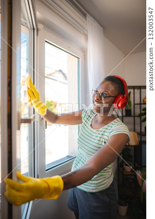 Happy housekeeper wearing headphones cleaning windows at home 132003547