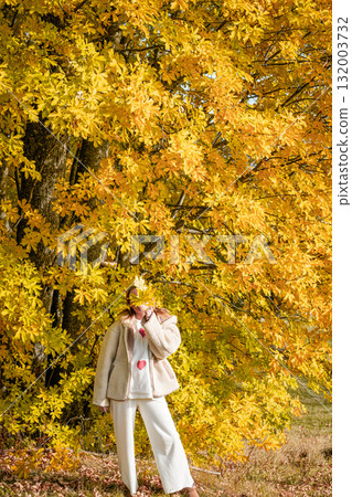 Scenic view of a woman standing under a yellow autumn tree, hiding her face behind fallen leaves. Warm fall colors create a peaceful seasonal atmosphere. 132003732