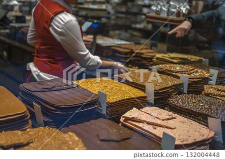 Stacks of artisan chocolate slabs displayed in a gourmet candy shop with seller assisting customers. Concept of chocolate indulgence, sweetness, handcrafted confectionery, sweet gift ideas 132004448