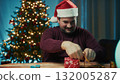 Young Man Wraps Christmas Gift On Table On Magical Night Against The Tree 132005287