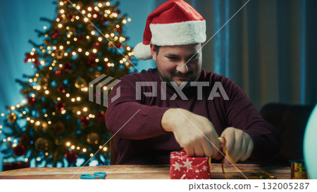 Young Man Wraps Christmas Gift On Table On Magical Night Against The Tree Young Man Wraps Christmas Gift On Table On Magical Night Against The Tree 132005287