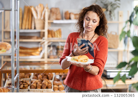 European female customer with phone takes pictures of burger croissant in disposable plate dish European female customer with phone takes pictures of burger croissant in disposable plate dish 132005365