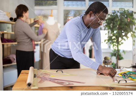 African man draw dress pattern of product on paper. Companion work in background with mannequin African man draw dress pattern of product on paper. Companion work in background with mannequin 132005574