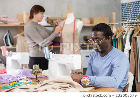 African American man tailor sews clothes, companion work in background with mannequin 132005579
