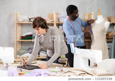 Woman tailor creates sketch of dress on tablet, man companion work in background with mannequin Woman tailor creates sketch of dress on tablet, man companion work in background with mannequin 132005599