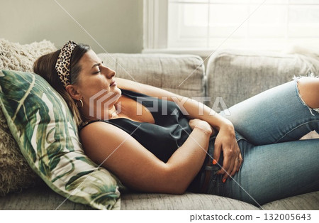 Period, menstrual pain and a woman on a sofa in the living room of her home with cramps of the abdomen. Stomach, digestion or menstruation with a young female person holding her belly in discomfort Period, menstrual pain and a woman on a sofa in the living room of her home with cramps of the abdomen. Stomach, digestion or menstruation with a young female person holding her belly in discomfort 132005643
