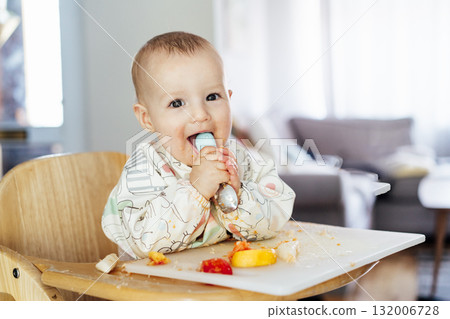 Happy baby learning to eat food with baby spoon by himself on high chair baby led weaning or blw. Infant self-feeding solid food, making mess, fine motor development. Kid enjoying healthy nutrition. Happy baby learning to eat food with baby spoon by himself on high chair baby led weaning or blw. Infant self-feeding solid food, making mess, fine motor development. Kid enjoying healthy nutrition. 132006728