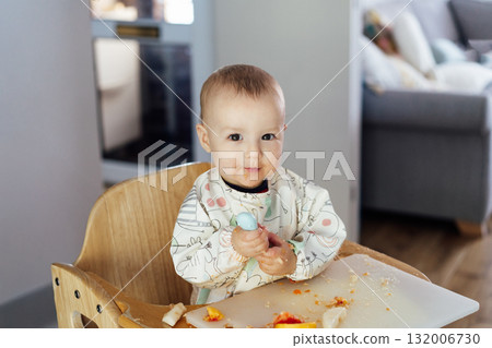 Cute baby learning to eat food with baby spoon by himself on high chair baby led weaning or blw. Infant self-feeding solid food, making mess, fine motor development. Kid enjoying healthy nutrition. 132006730