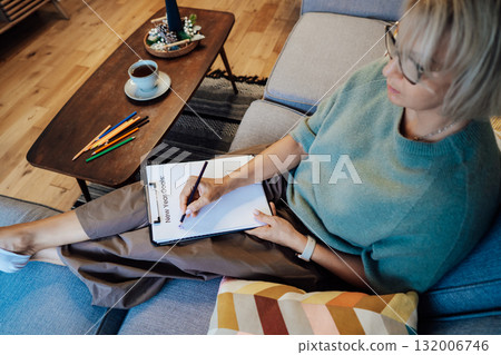Middle aged woman writing New year's goals on note paper. New year resolutions list, cup of tea on table. Goals, resolutions, plans, action, checklist concept. New Year template, copy space Middle aged woman writing New year's goals on note paper. New year resolutions list, cup of tea on table. Goals, resolutions, plans, action, checklist concept. New Year template, copy space 132006746