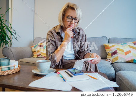 Middle aged woman reviewing expenses receipts while sitting on couch with coffee and phone in cozy living space. Managing personal finances with mobile app . Checking financial balance, budgeting. Middle aged woman reviewing expenses receipts while sitting on couch with coffee and phone in cozy living space. Managing personal finances with mobile app . Checking financial balance, budgeting. 132006749