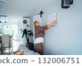 Side view middle aged woman putting painting picture in frame on the wall. Casual lady taking care of coziness in her new stylish apartment. Mature female decorating room. Moving and renovate house 132006751