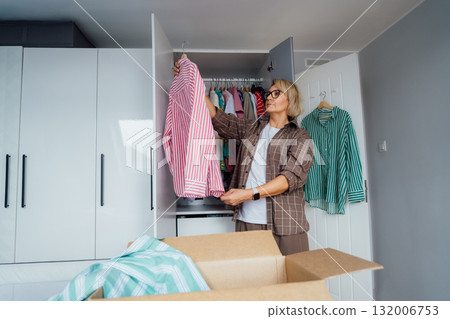 Middle aged woman selecting clothes from wardrobe for donating to Charity shop. Decluttering, Sorting clothes and Cleaning Up. Reuse, second-hand concept. Conscious consumer, sustainable lifestyle. Middle aged woman selecting clothes from wardrobe for donating to Charity shop. Decluttering, Sorting clothes and Cleaning Up. Reuse, second-hand concept. Conscious consumer, sustainable lifestyle. 132006753