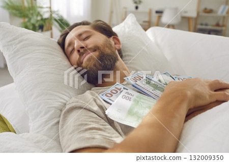 Smiling young man sleeping on bed with a bunch of cash dollars keeping his money savings at home. 132009350
