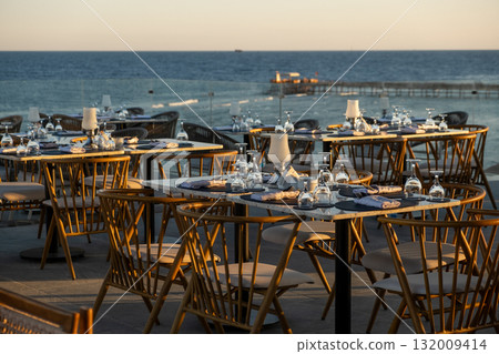 Seaside restaurant tables set for an exquisite dining experience, featuring glassware and a sunset-lit ocean backdrop 132009414