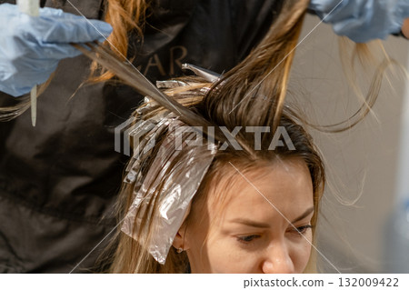 A client is having her hair colored while seated in a salon, with a stylist applying dye and using foils for highlights. A client is having her hair colored while seated in a salon, with a stylist applying dye and using foils for highlights. 132009422