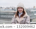 A young woman stands outdoors in a warm jacket and knitted hat, smiling softly while the city skyline looms in the background on a cloudy day. 132009462