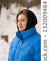 A woman wearing a blue winter coat and black headscarf looks thoughtfully into the distance against a snowy backdrop filled with trees. 132009464