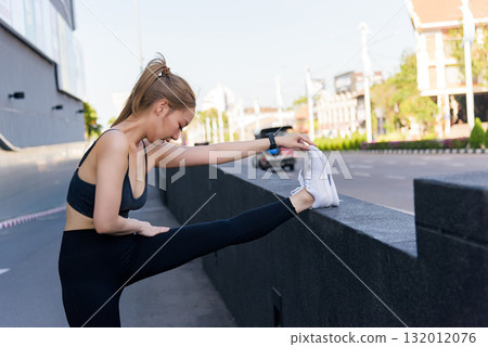 Sport woman performs outdoor exercise, stretching leg for fit workout preparation. Young healthy athletic person trains vigorously, enjoying urban street sport activity, pursuing active lifestyle. 132012076