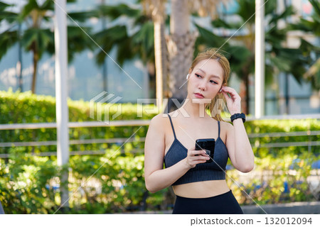 Fit sport woman outdoor adjusts earbud, holds phone, prepares workout, begins pre workout stretch routine, ready to stretch muscles, embracing healthy active lifestyle, finding fitness inspiration. Fit sport woman outdoor adjusts earbud, holds phone, prepares workout, begins pre workout stretch routine, ready to stretch muscles, embracing healthy active lifestyle, finding fitness inspiration. 132012094