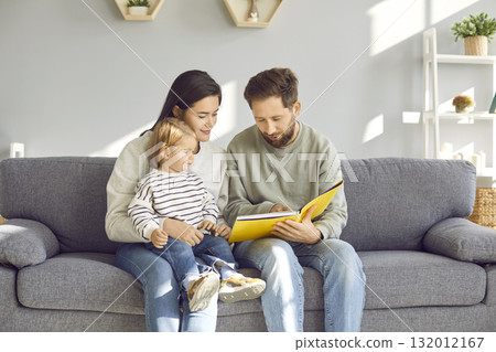 Happy family sitting on sofa reading a book together in their living room 132012167