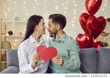 Attractive happy couple holding red heart together sitting on couch at home Attractive happy couple holding red heart together sitting on couch at home 132012172