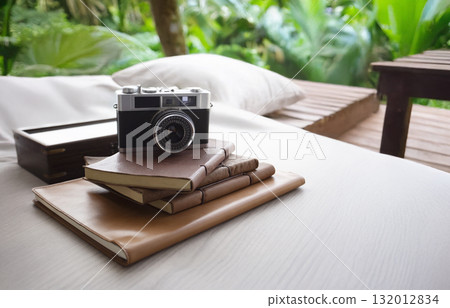 Vintage camera and leather-bound journals resting on a daybed in a lush, tropical setting. Perfect moment for travel, reflection, and creative escape 132012834