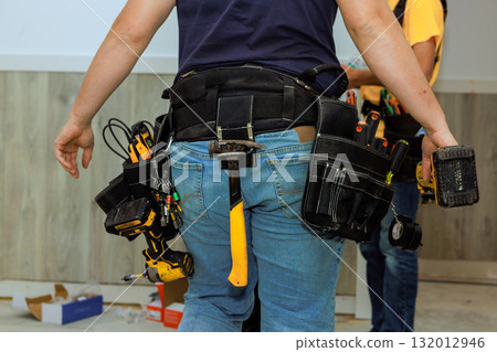 Worker wearing tool belt walks in construction area, ready for upcoming renovation work, surrounded by materials. 132012946