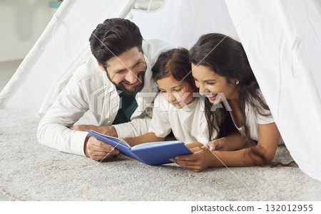 Happy mother and father read story book to daughter, lying on carpet in hygge tent at home playroom Happy mother and father read story book to daughter, lying on carpet in hygge tent at home playroom 132012955