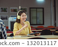 Happy young Asian student wearing headphones and using a tablet, enjoying online lessons, studying in the library. 132013117