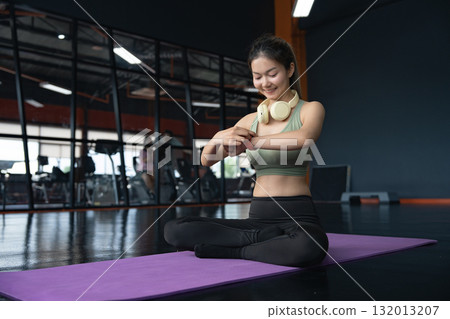 Young Asian woman in sportswear checking her workout results on a smart watch while resting at the gym. 132013207