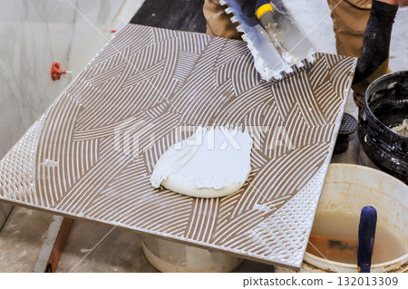 Worker spreads adhesive onto large tile, preparing it for installation on floor. Worker spreads adhesive onto large tile, preparing it for installation on floor. 132013309