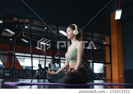 Young woman sitting on the floor on mats and practicing yoga in lotus position with eyes closed in a gym. 132013352