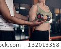 Young asian woman doing exercises with dumbbells in a gym while her trainer is supporting her. 132013358