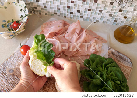 Anonymous person makes sandwich with sausage, lettuce, and mortadella, homemade mayonnaise in kitchen. Italian cuisine, high-quality farm-grown ingredients. 132015528