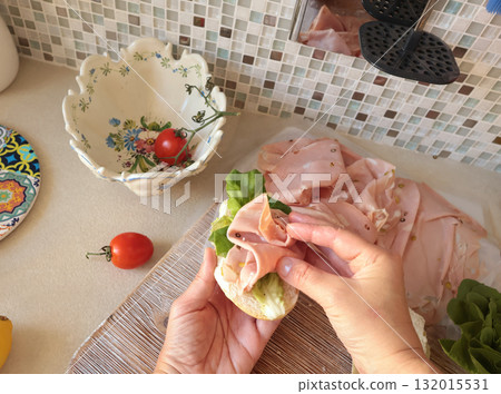 Anonymous person makes sandwich with sausage, lettuce, and mortadella, homemade mayonnaise in kitchen. Italian cuisine, high-quality farm-grown ingredients. Anonymous person makes sandwich with sausage, lettuce, and mortadella, homemade mayonnaise in kitchen. Italian cuisine, high-quality farm-grown ingredients. 132015531