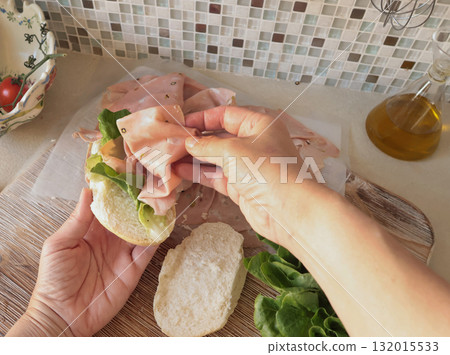 Anonymous person makes sandwich with sausage, lettuce, and mortadella, homemade mayonnaise in kitchen. Italian cuisine, high-quality farm-grown ingredients. 132015533