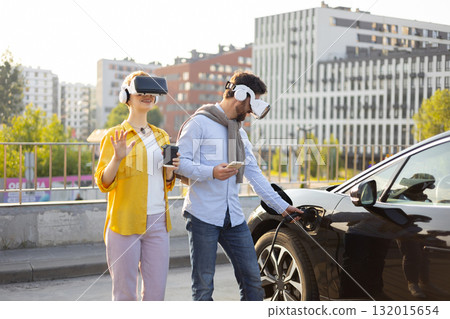 A couple wearing VR headsets while one charges an electric vehicle in an urban setting. A couple wearing VR headsets while one charges an electric vehicle in an urban setting. 132015654