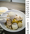 A fishball is picked up by a pair of chopsticks in the foreground against the background of a bowl of minced meat noodle 132016721