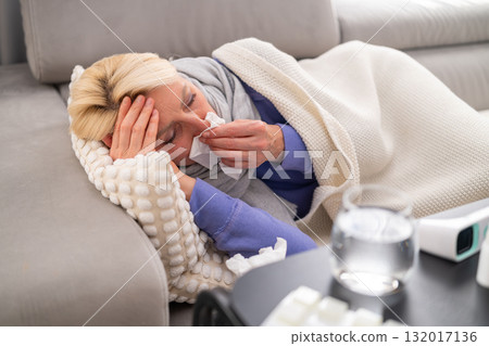 Blonde woman lying on couch with runny nose, covered with blanket, looking tired. Pills, thermometer, and tissues in the foreground. Concept of flu, illness, and home recovery. Blonde woman lying on couch with runny nose, covered with blanket, looking tired. Pills, thermometer, and tissues in the foreground. Concept of flu, illness, and home recovery. 132017136