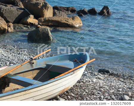 Old white and blue wooden boat with oars on shore near rocks at sunset near sea. Peace, quiet, and relaxation. Design background. Old white and blue wooden boat with oars on shore near rocks at sunset near sea. Peace, quiet, and relaxation. Design background. 132017609