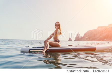 Woman Paddleboarding Ocean Coastline - Young woman in black bikini relaxing on paddleboard in ocean with coastal cliffs in background. Woman Paddleboarding Ocean Coastline - Young woman in black bikini relaxing on paddleboard in ocean with coastal cliffs in background. 132020661
