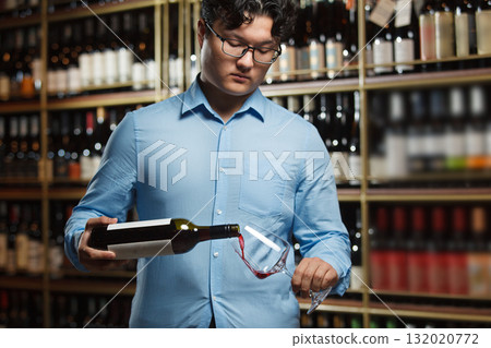 Expert Asian Sommelier Pouring Red Wine in Glass Amidst an Extensive Wine Cellar Collection Expert Asian Sommelier Pouring Red Wine in Glass Amidst an Extensive Wine Cellar Collection 132020772