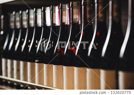 Row of Red Wine Bottles Lined Up on Wooden Shelves in Cozy Cellar 132020779