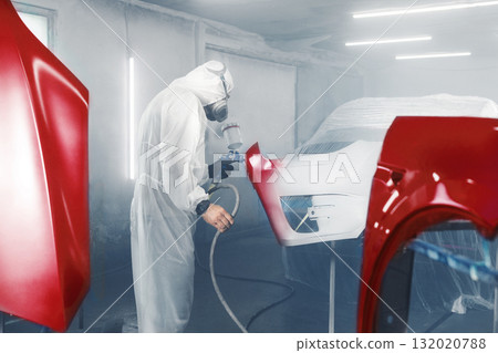 Car paint worker spraying red paint to car body element using spray gun in vehicle workshop chamber. Complex restoration of automobile body.  132020788