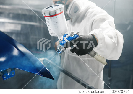 Male automobile repairman painter in protective workwear and respirator painting part of car body in paint chamber 132020795